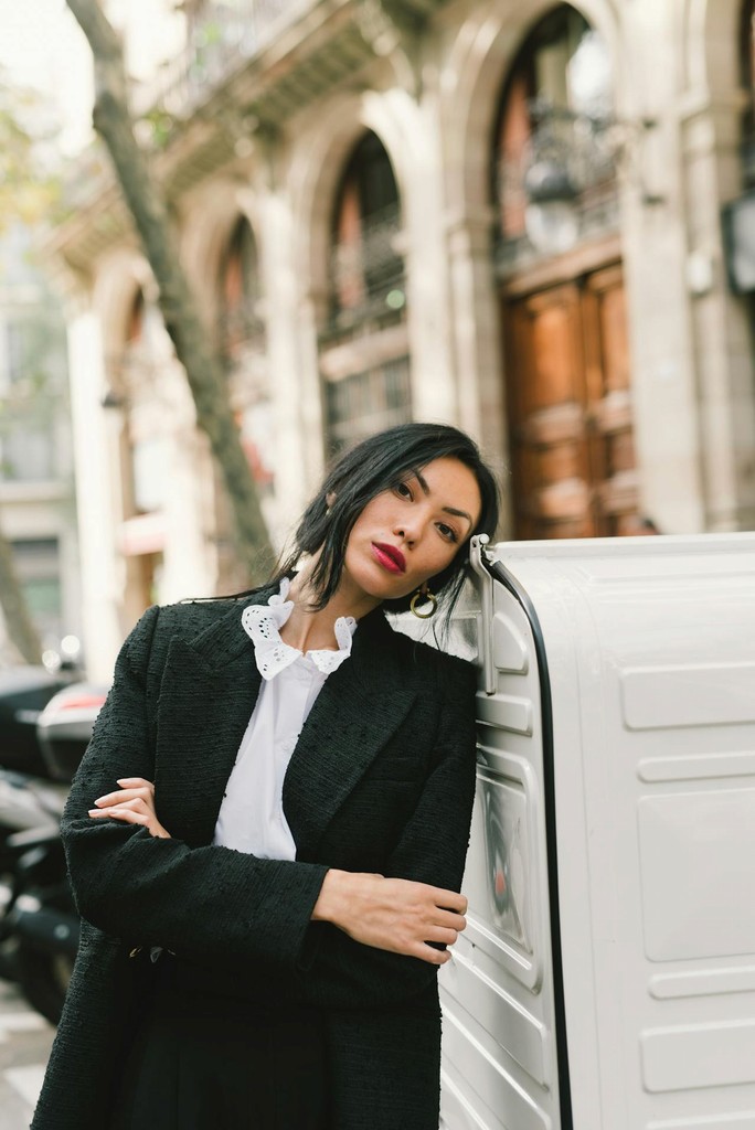 Woman in a black blazer and ruffled shirt leans on a white delivery box in an urban street with historic buildings.