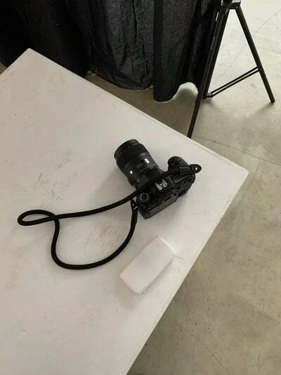 Black camera with a rope strap and white object on a white table. A tripod and black backdrop are in the background.